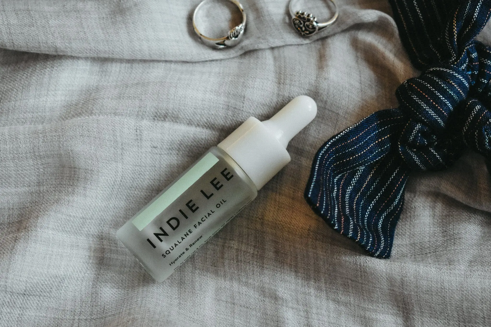 Why-Use-Facial-Oil-Other-Facial-Oil-FAQ4 - Cats & Coffee Indie Lee's Squalane Facial Oil on a blue backdrop, surrounded by a ribbon hair bow and rings