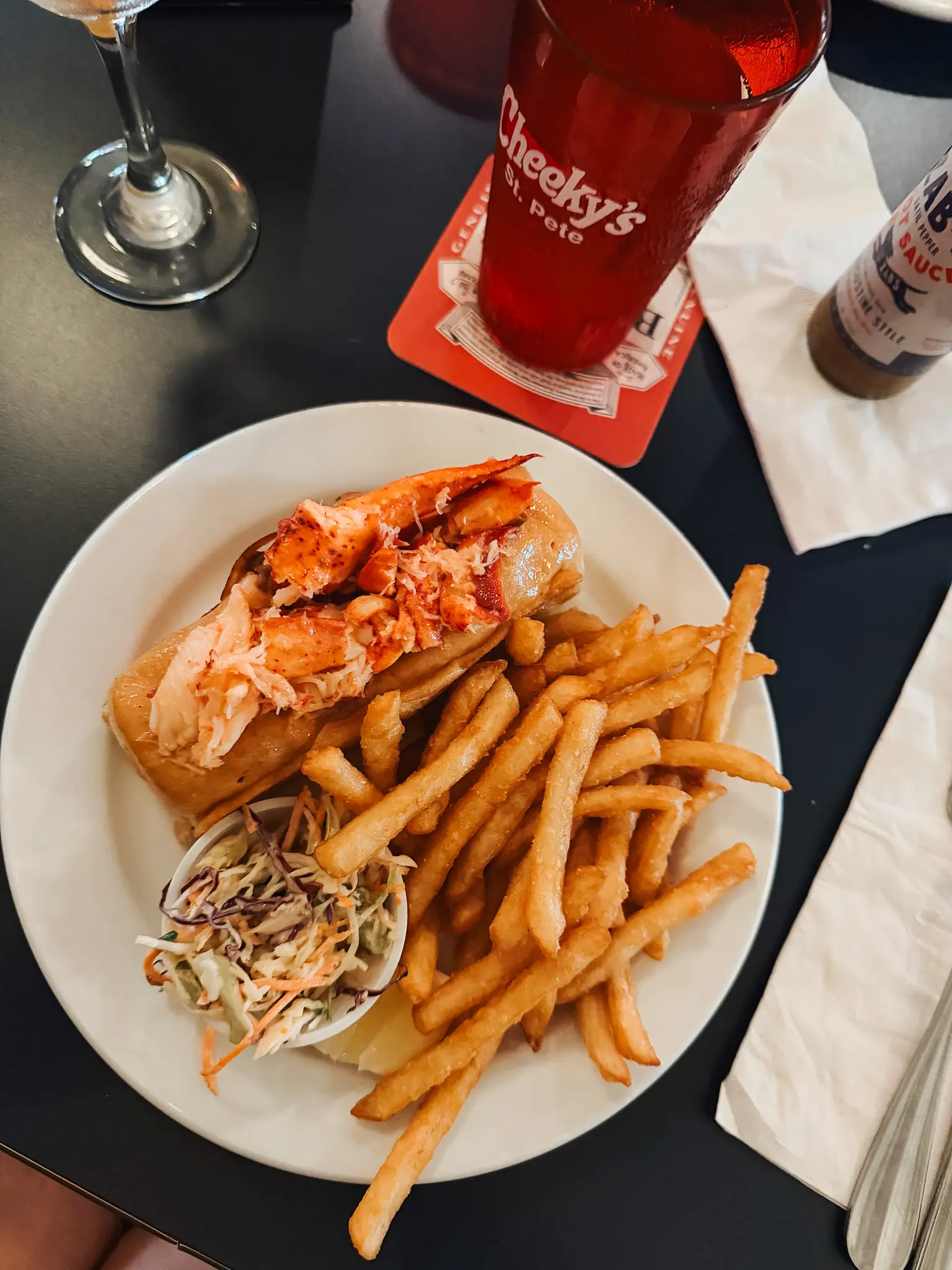 Cheekys St Pete Restaurant Clam Bar 2 - Cats & Coffee Lobster roll with fries and coleslaw on a white plate; a red drink and napkin on a dark table.