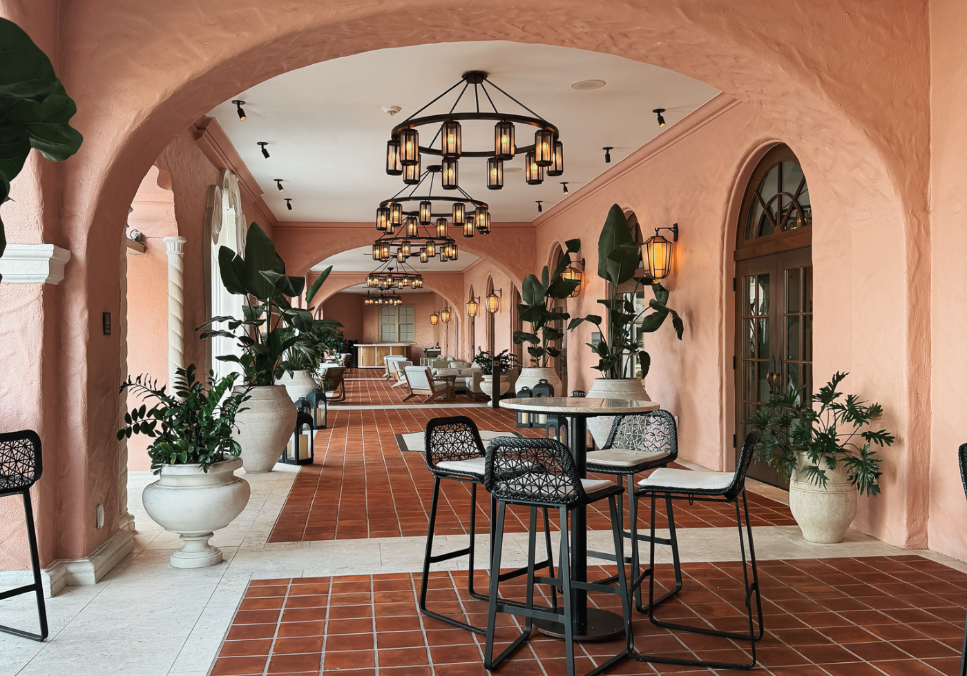 The Best Places to Stay in Tampa Florida - Cats & Coffee Peach-colored arched veranda with terracotta tiles, large potted plants, and black metal high tables with woven chairs under lanterns.