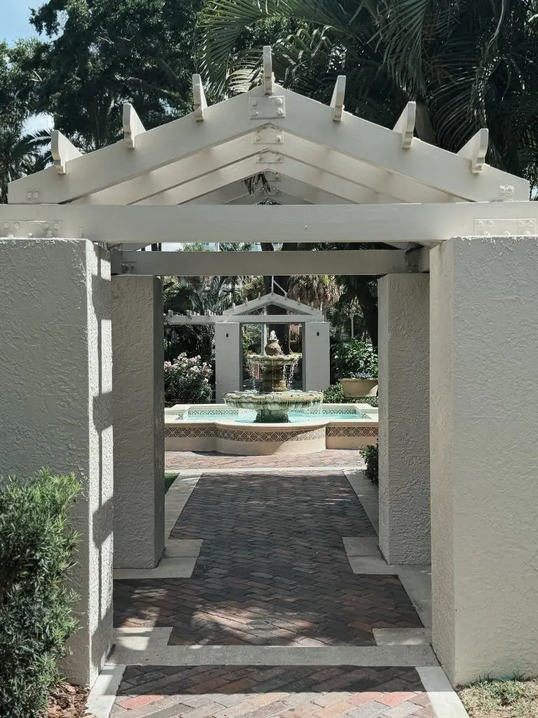 The Vinoy St Pete - Where to Stay in Tampa Bay - 1 - Cats & Coffee White-painted wooden pergola arches over a brick-paved walkway leading to a tiered fountain in a manicured garden.