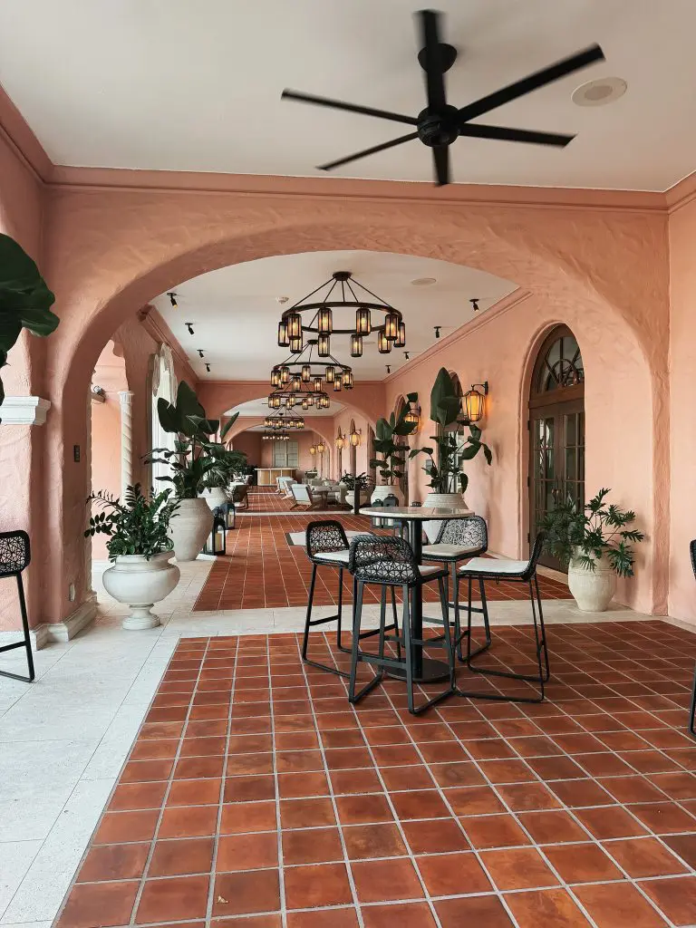 The Vinoy St Pete - Where to Stay in Tampa Bay - 2 - Cats & Coffee Pink arched corridor with terracotta tile floor, large potted plants, and black lantern chandeliers along the hallway.