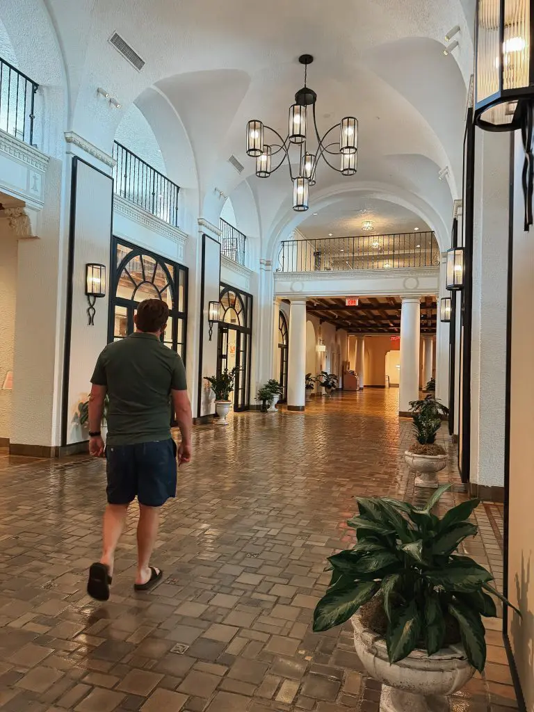 The Vinoy St Pete - Where to Stay in Tampa Bay - 4 - Cats & Coffee Man in a green shirt and shorts walks away down a tiled, arched indoor hallway with lantern-style lights and potted plants along storefronts.