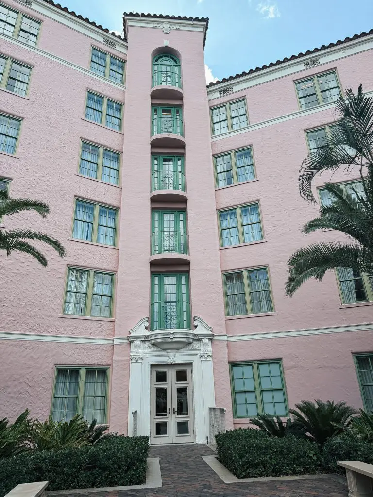 The Vinoy St Pete - Where to Stay in Tampa Bay - 7 - Cats & Coffee Pink stucco building with teal-green doors and windows, central vertical balconies, and decorative entrance in a courtyard with hedges and palm branches nearby