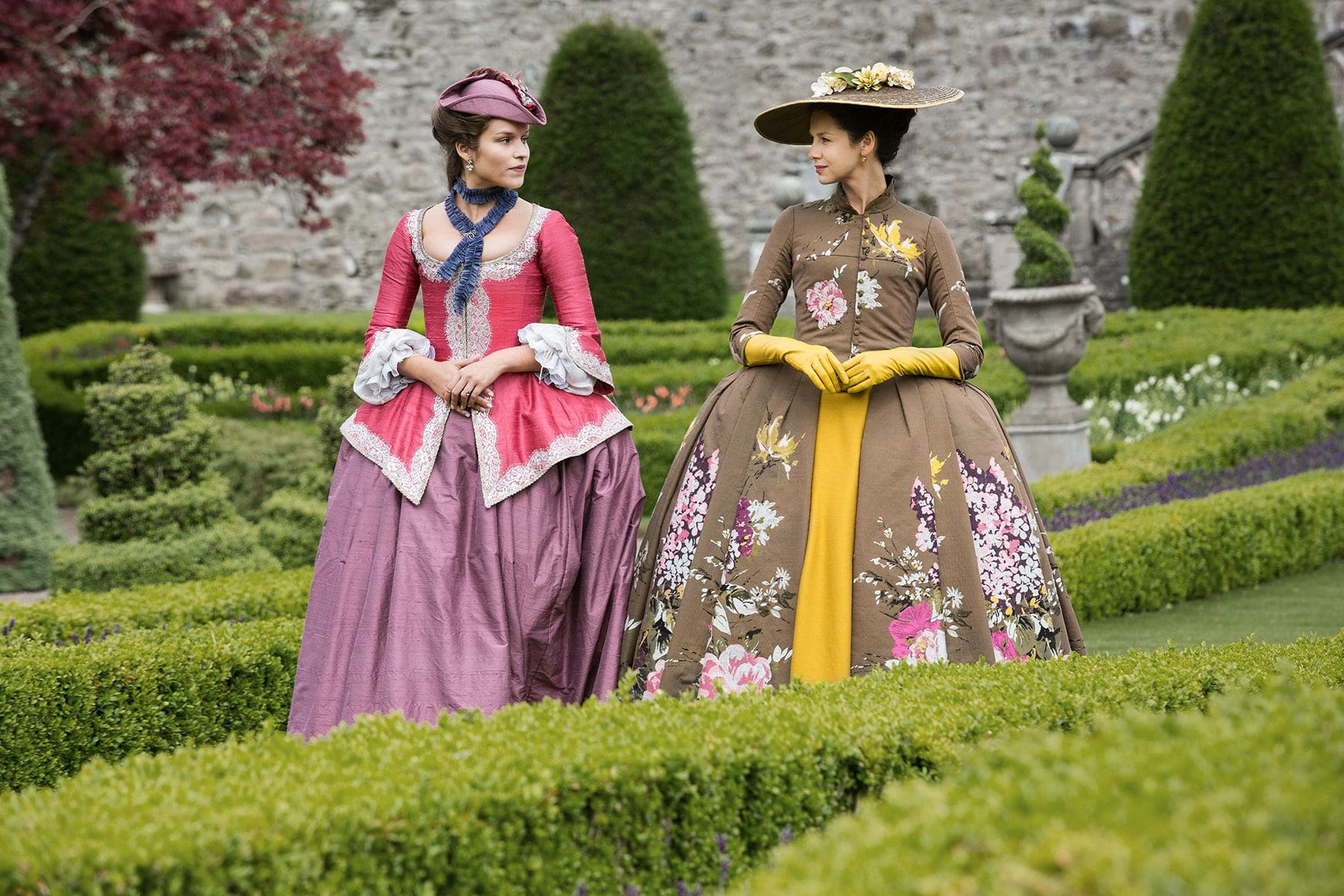 Annalise de Marillac from Outlander Season 2 in a pink and purple gown with blue collar detailing; walking with Claire Fraser in a brown and yellow floral gown in the gardens of Versailles